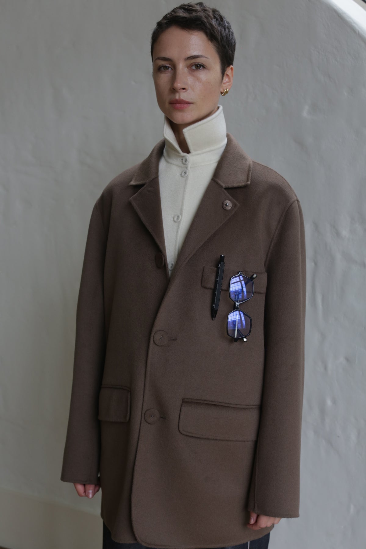 A person with short hair wears the Sculptor's Jacket in Taupe over a buttoned white turtleneck. Sunglasses are clipped to the jacket pocket, catching light. The background is a simple, light-colored wall.