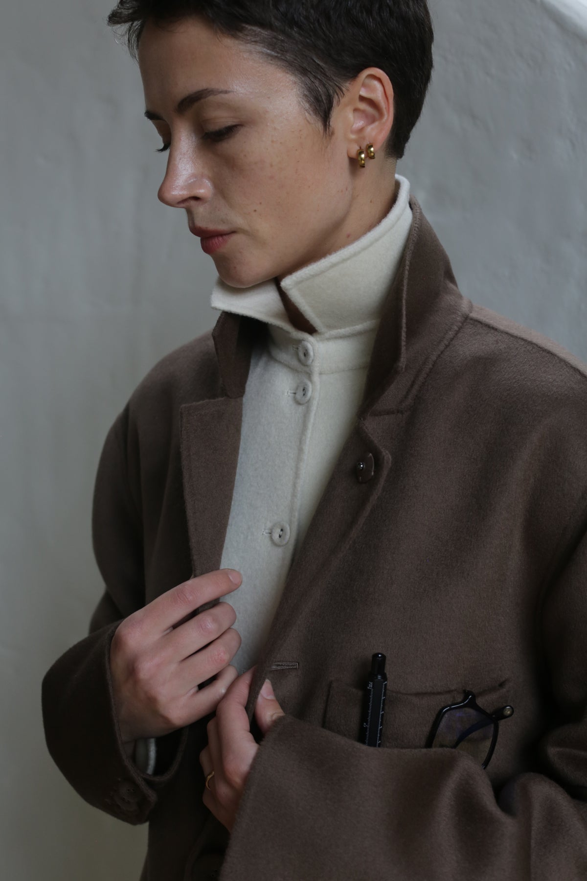 A person with short dark hair wears the Sculptor's Jacket | Taupe over a cream buttoned turtleneck, looking down thoughtfully. Glasses and a pen peek from the chest pocket as they adjust their coat.