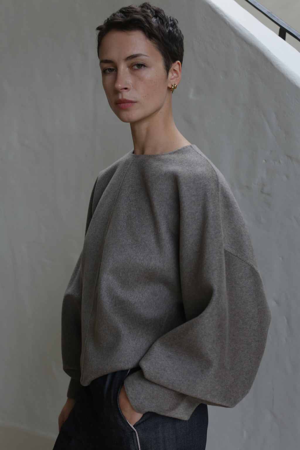 A person with short dark hair wearing the Cocoon | Undyed Natural, a loose long-sleeved top, stands against a light-textured wall and looks at the camera with a neutral expression.