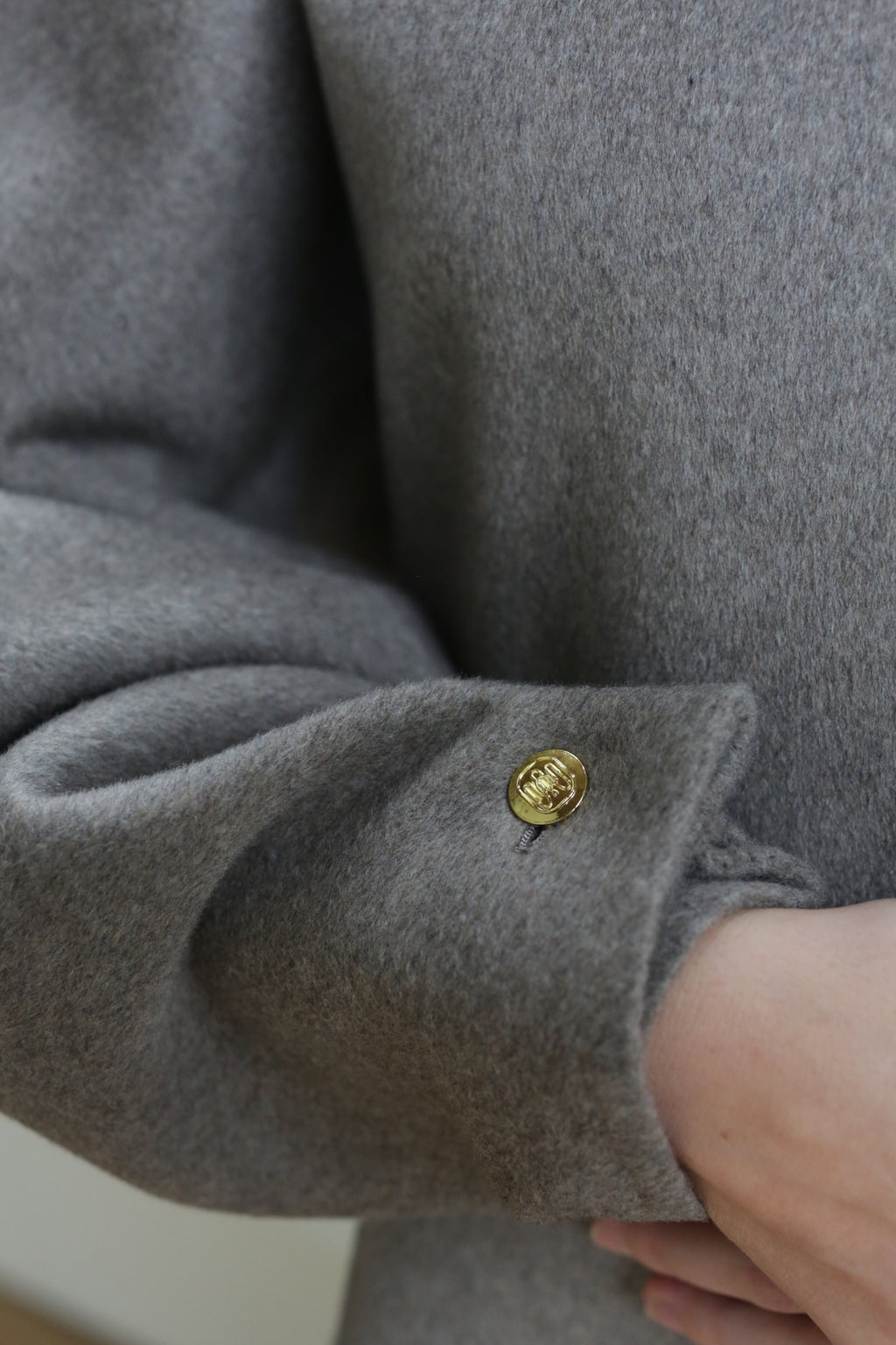 A close-up of a hand partially tucked into the sleeve of the Cocoon | Undyed Natural, a gray wool coat with a shiny gold button near the cuff.