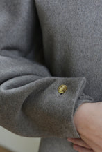 Load image into Gallery viewer, A close-up of a hand partially tucked into the sleeve of the Cocoon | Undyed Natural, a gray wool coat with a shiny gold button near the cuff.