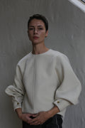 A person with short dark hair stands against a light grey wall, wearing the Cocoon | Undyed—a loose-fitting, long-sleeved white top featuring gold buttoned cuffs. Their expression is calm and neutral.