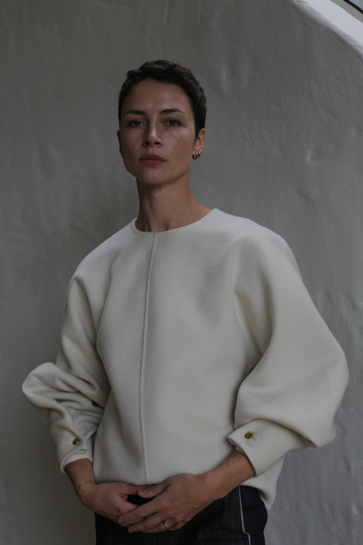 A person with short dark hair stands against a light grey wall, wearing the Cocoon | Undyed—a loose-fitting, long-sleeved white top featuring gold buttoned cuffs. Their expression is calm and neutral.