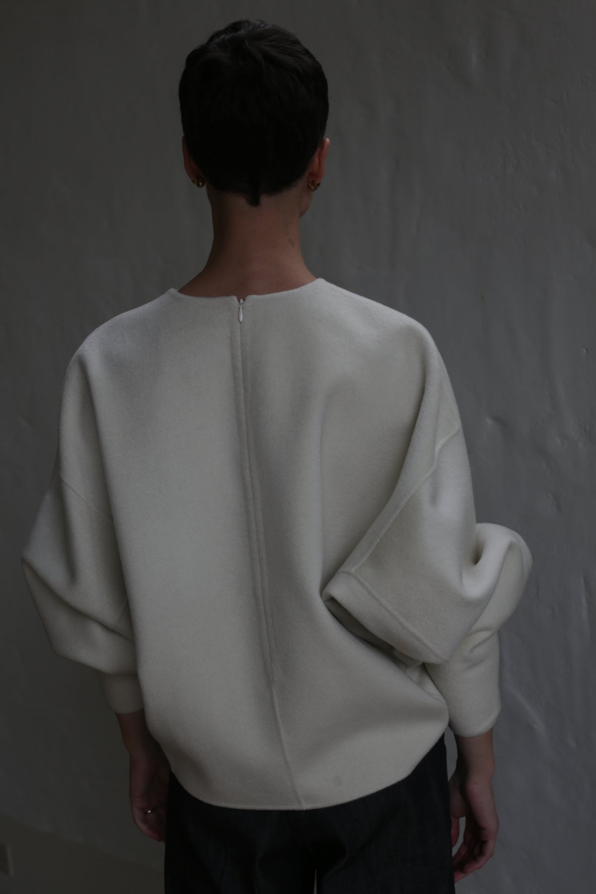 A person with short dark hair stands with their back to the camera, wearing the Cocoon | Undyed—an off-white, loose-fitting top with wide sleeves—against a plain textured gray wall.