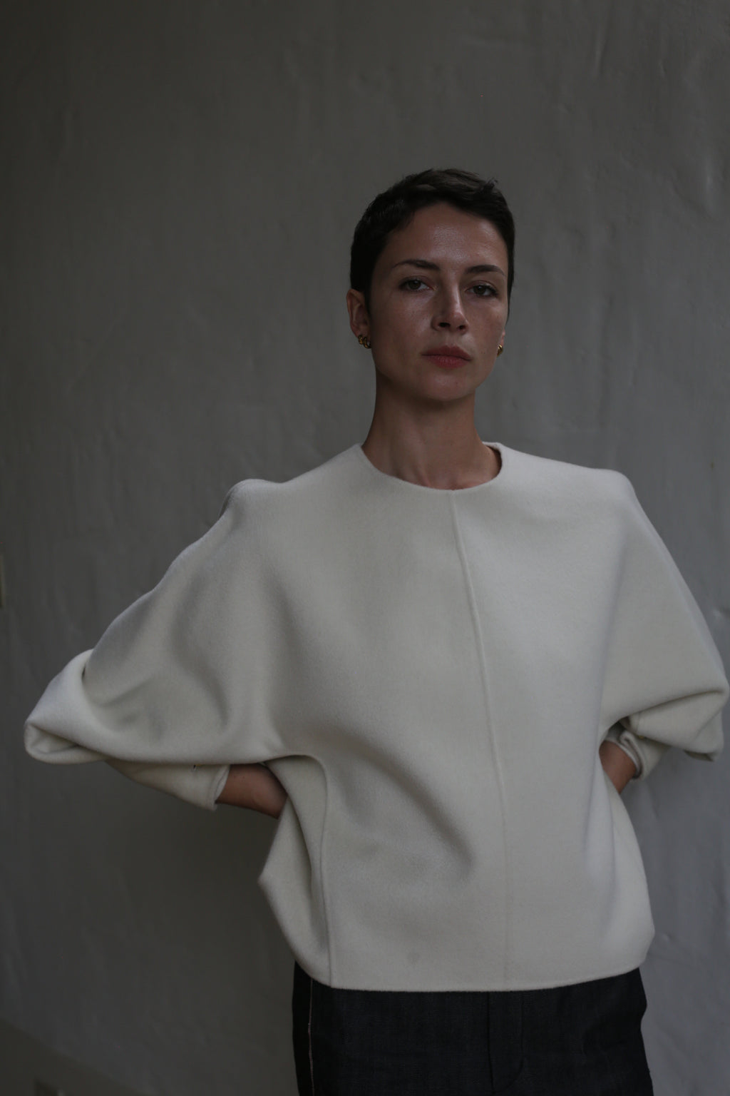 A person with short dark hair stands against a gray textured wall, wearing the Cocoon | Undyed wide-sleeved top. With hands behind their back, they look calmly at the camera under soft, subdued lighting.