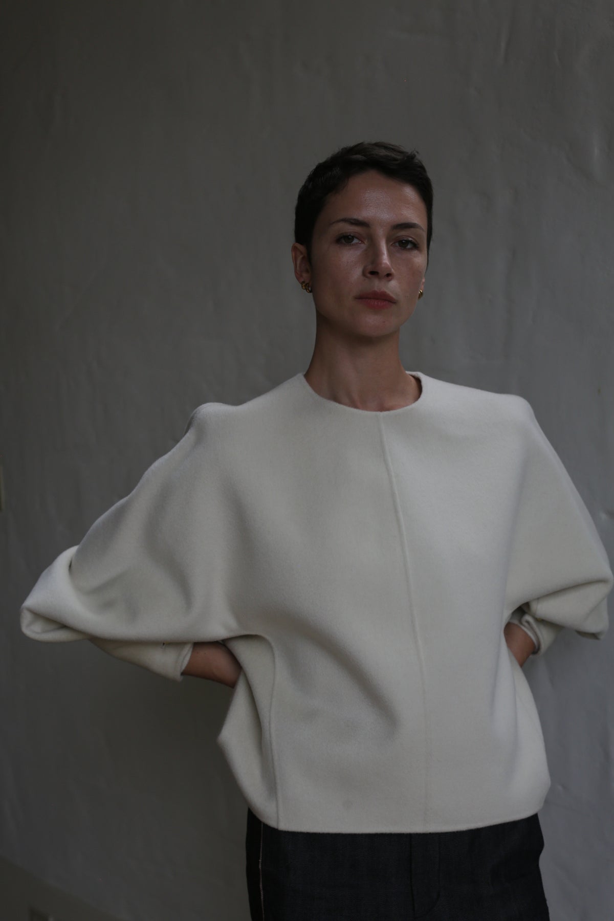 A person with short dark hair stands against a gray textured wall, wearing the Cocoon | Undyed wide-sleeved top. With hands behind their back, they look calmly at the camera under soft, subdued lighting.