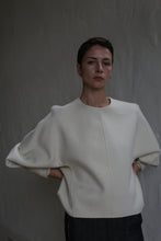 Load image into Gallery viewer, A person with short dark hair stands against a gray textured wall, wearing the Cocoon | Undyed wide-sleeved top. With hands behind their back, they look calmly at the camera under soft, subdued lighting.