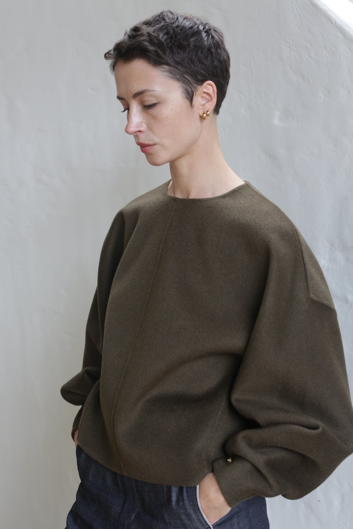 A person with short dark hair and gold earrings stands against a light wall, looking down with hands in pockets, wearing the Cocoon | Double Face Cashmere Olive luxury oversized unisex top.