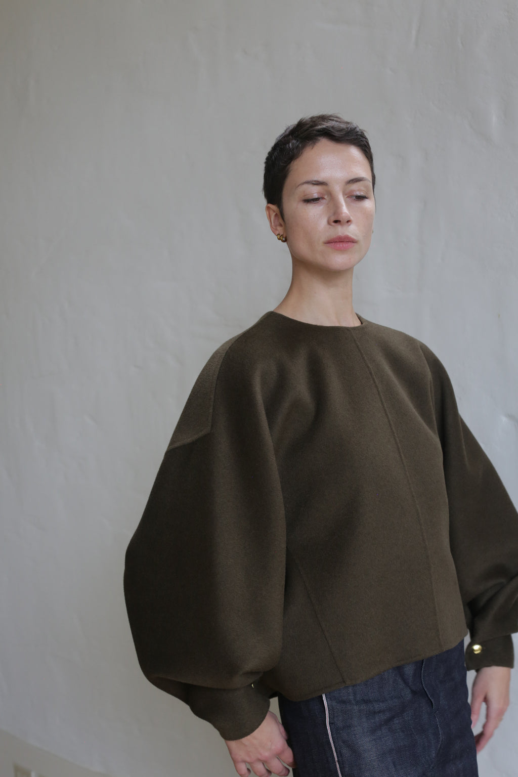 A person with short dark hair wears the Cocoon Double Face Cashmere Olive unisex top with wide sleeves and dark pants, standing against a light textured wall. They have gold stud earrings and a calm, neutral expression—understated luxury.