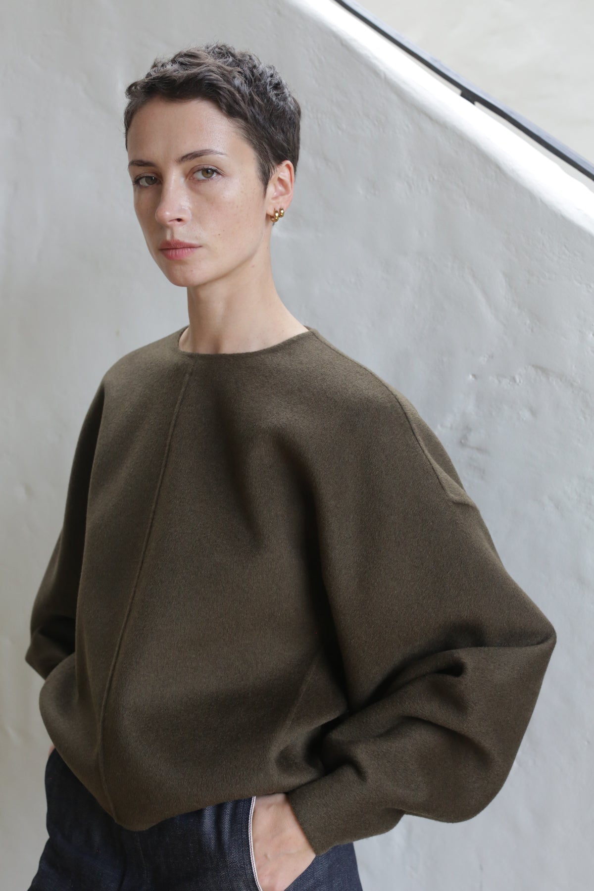A person with short dark hair wears the Cocoon | Double Face Cashmere Olive luxury unisex top and dark pants, standing with one hand in their pocket before a white textured wall, gazing slightly to the side.