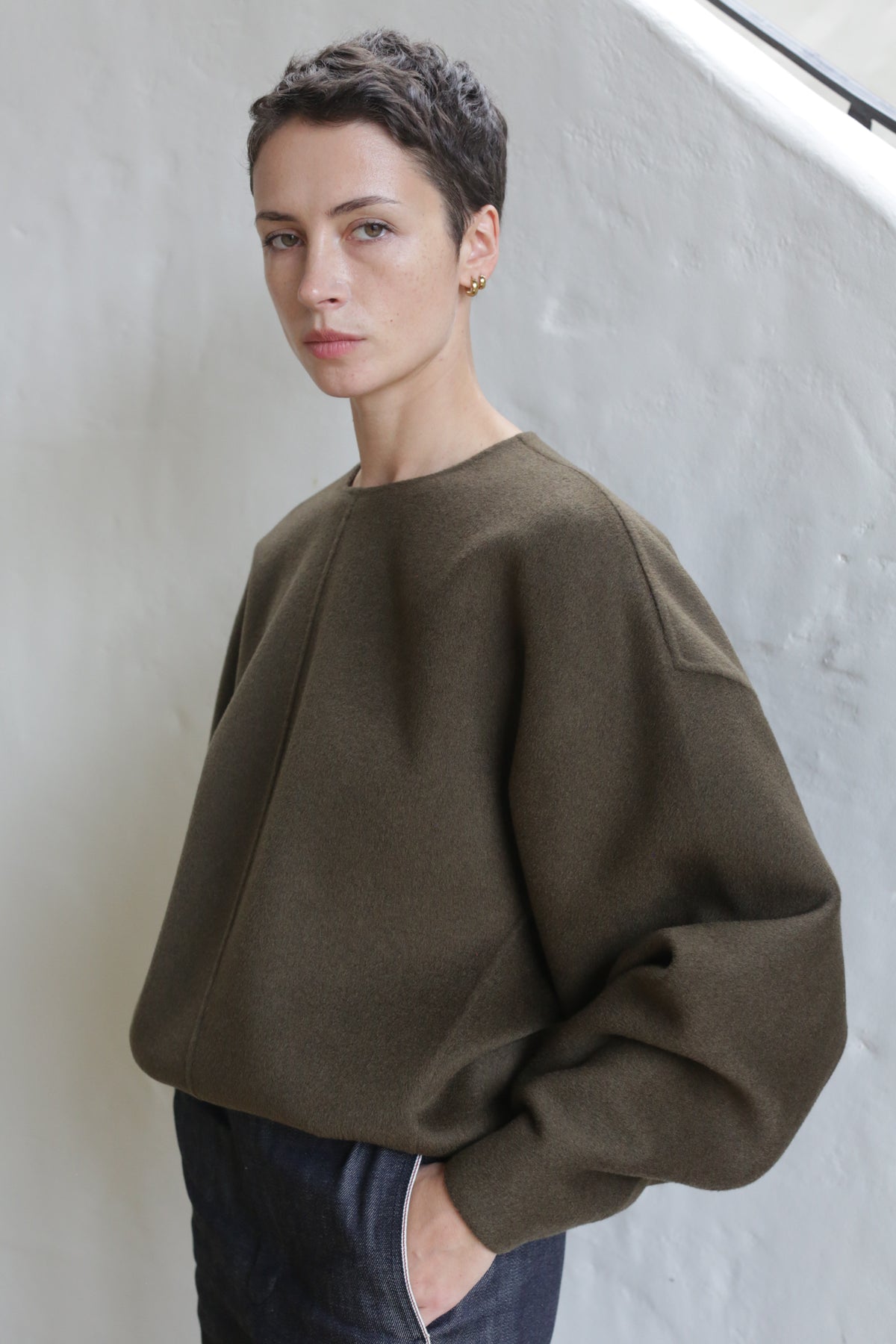 A person with short brown hair wears the Cocoon | Double Face Cashmere Olive top—an unisex, loose, olive-brown luxury piece—with dark pants, standing against a white curved wall and looking at the camera, one hand in their pocket.