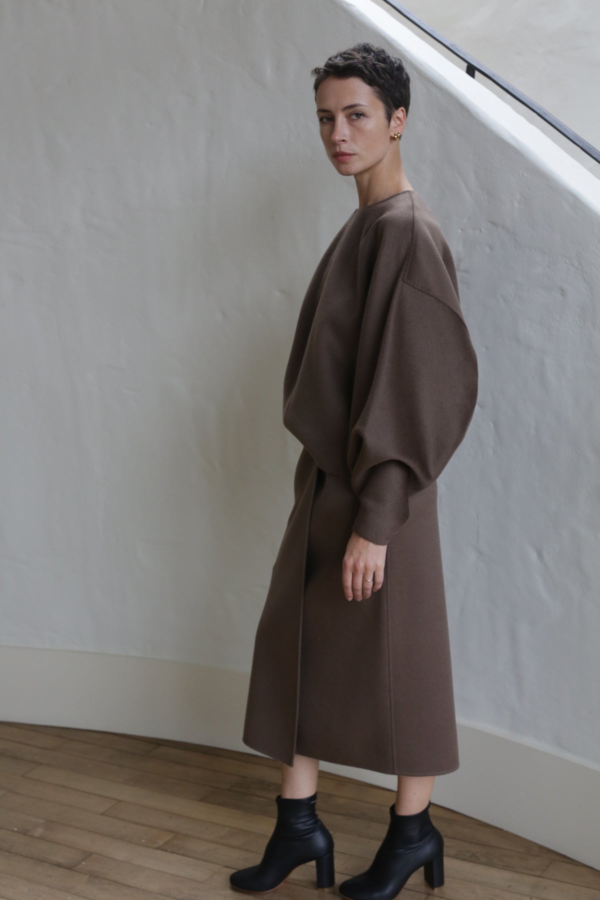 A person with short hair wears Cocoon | Taupe, a loose long-sleeved dress with voluminous sleeves, paired with black ankle boots, standing indoors against a white textured wall and wooden floor.