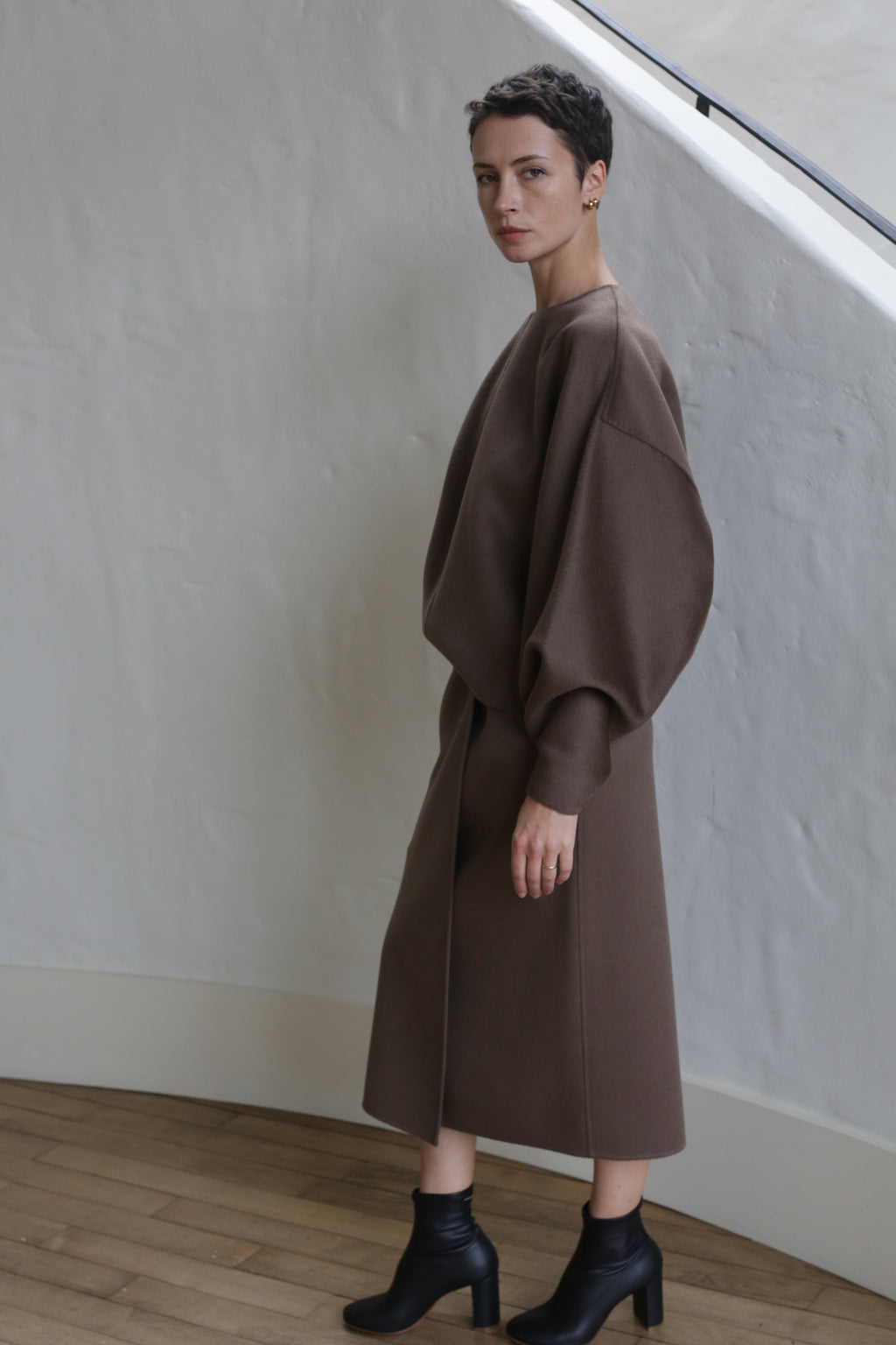 A person with short dark hair wears the Wrap Skirt in Taupe and black ankle boots, standing indoors against a white wall and looking at the camera.