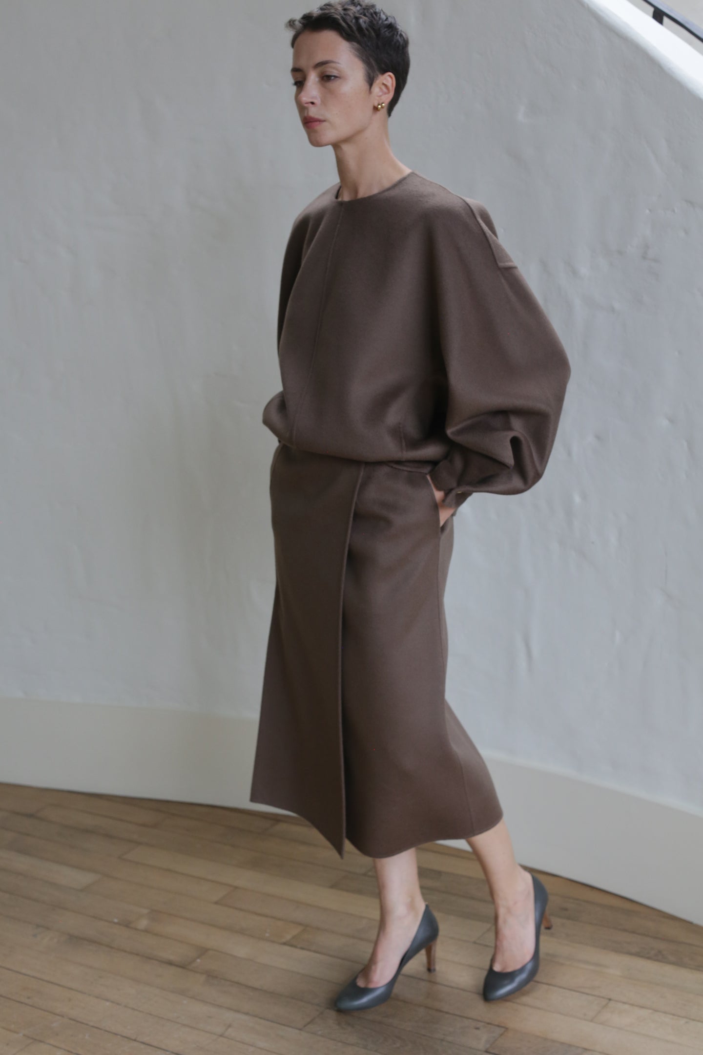 A person with short hair stands indoors against a light-colored textured wall, wearing the Wrap Skirt in Taupe and a loose-fitting, long-sleeved brown top, paired with dark heeled shoes.