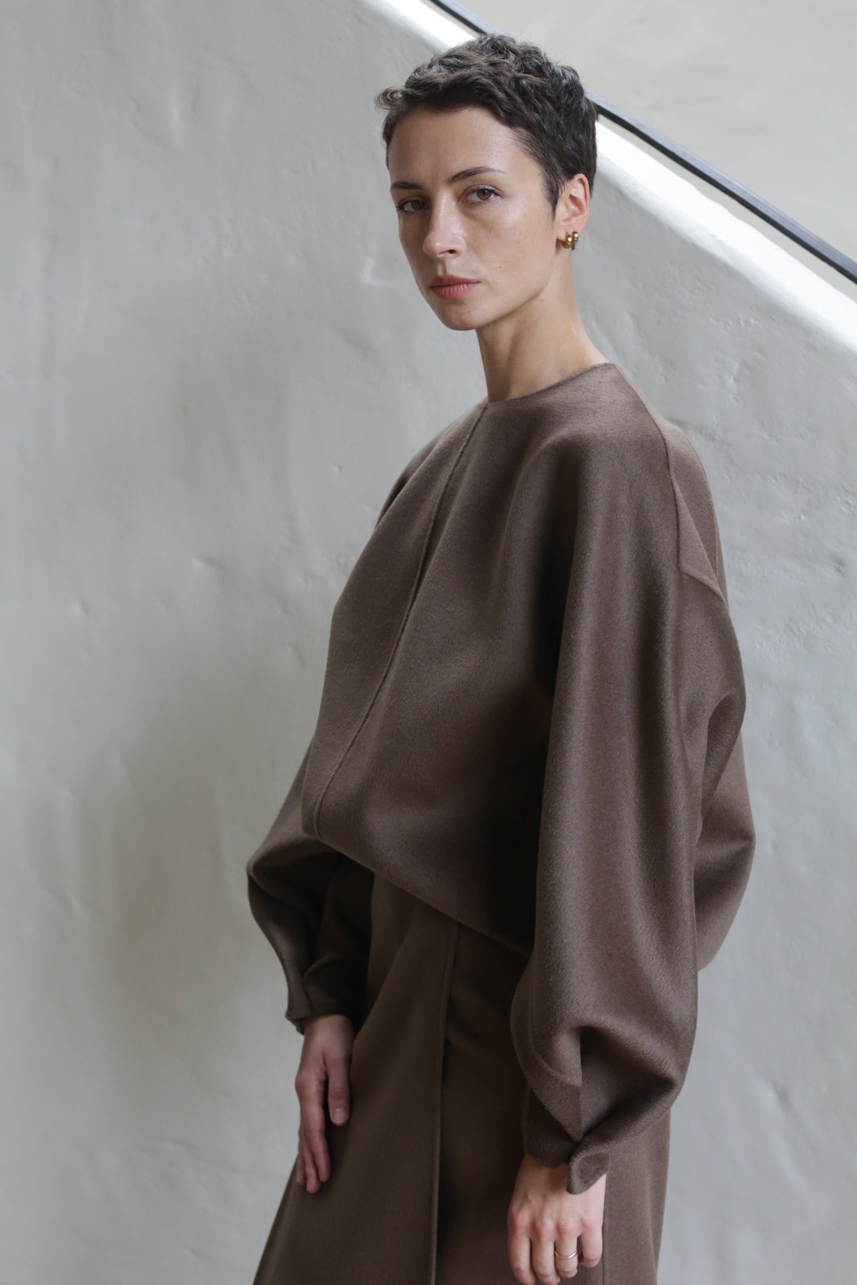 A person with short dark hair wears the Cocoon | Taupe set—loose-fitting, long-sleeved top and matching skirt—standing against a light-colored textured wall and looking slightly to the side with a neutral expression.