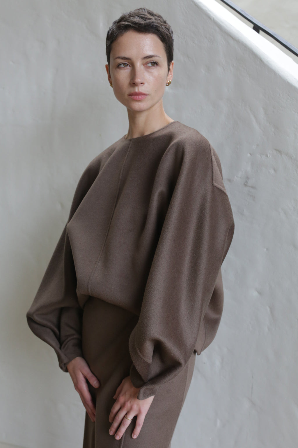 A person with short dark hair models the Cocoon | Taupe, a loose-fitting top with voluminous sleeves, standing against a light-colored textured wall and gazing thoughtfully to the side.