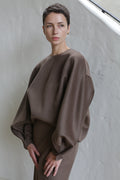 A person with short dark hair models the Cocoon | Taupe, a loose-fitting top with voluminous sleeves, standing against a light-colored textured wall and gazing thoughtfully to the side.