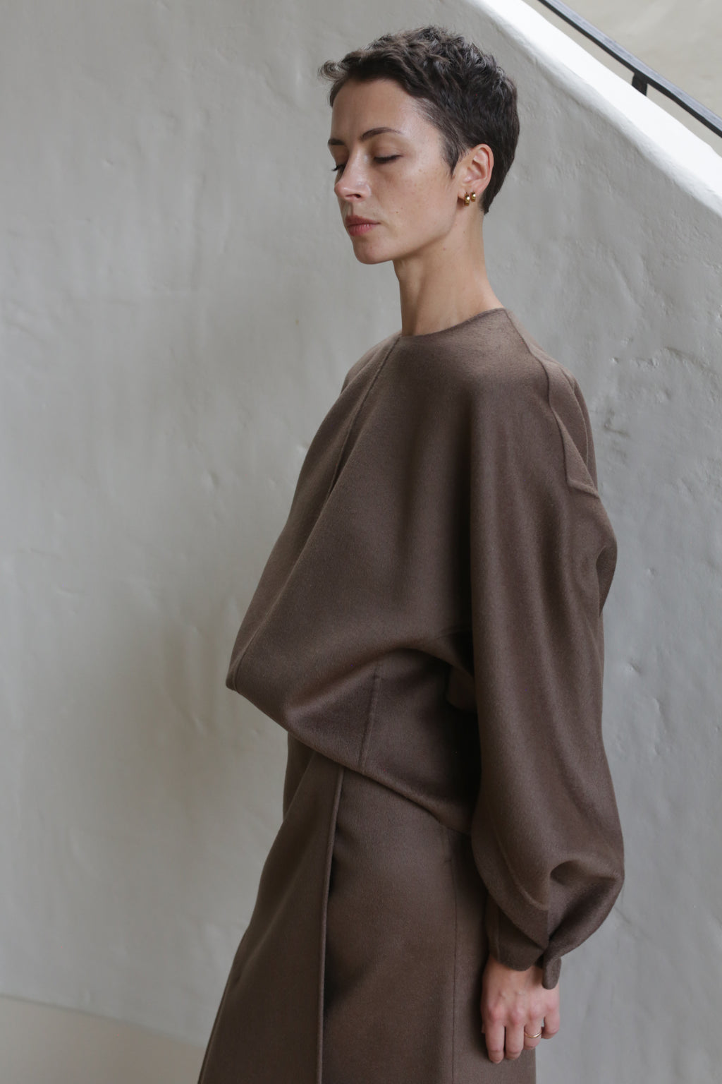 A person with short, dark hair stands in profile, eyes closed, wearing the Lan Jaenicke Cocoon Double Face Cashmere Taupe top and a matching brown skirt against a light-colored wall.