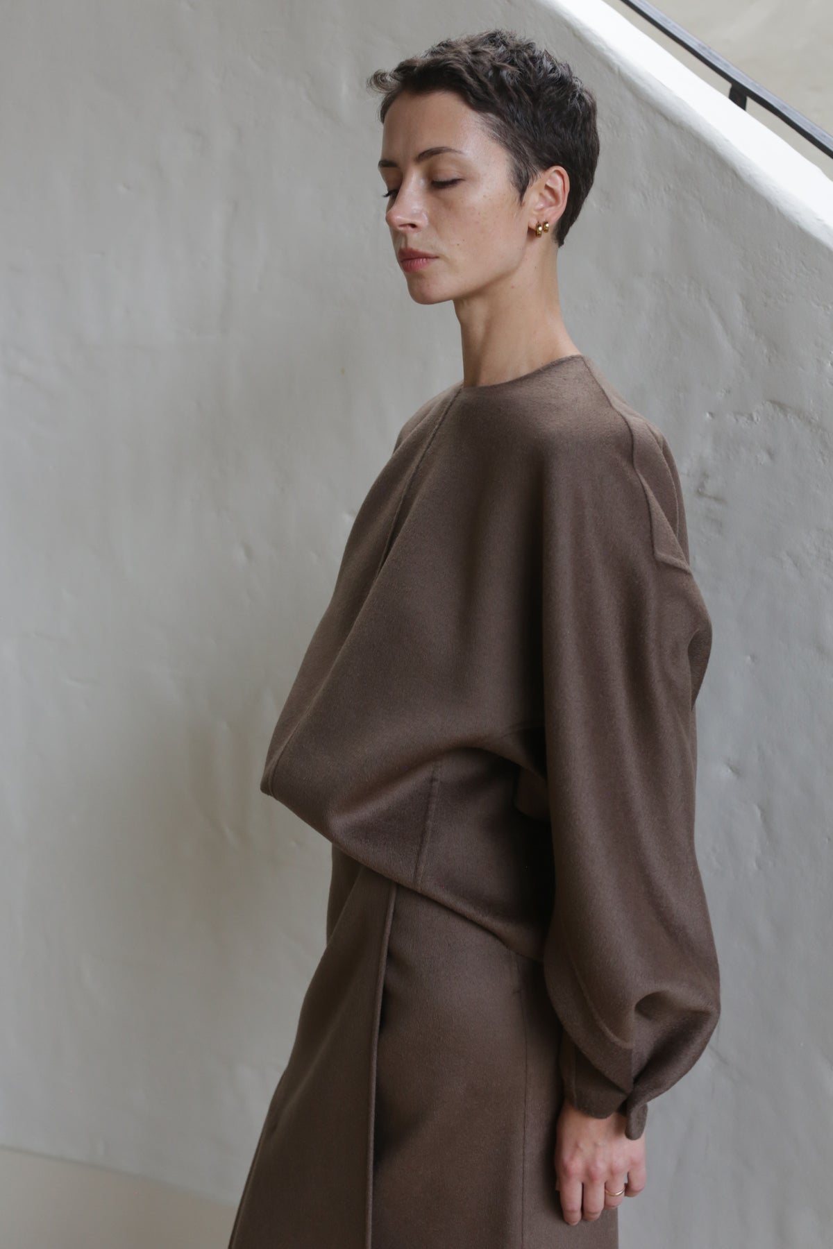 A person with short, dark hair stands in profile, eyes closed, wearing the Lan Jaenicke Cocoon Double Face Cashmere Taupe top and a matching brown skirt against a light-colored wall.