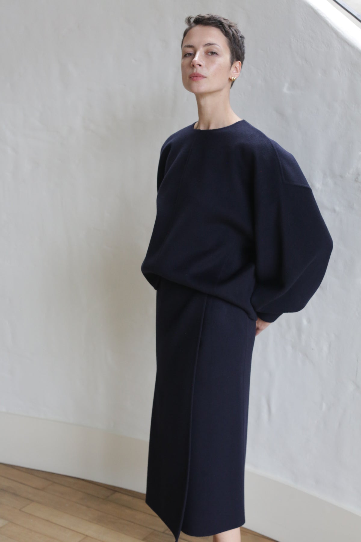 A person with short hair stands indoors against a white textured wall, wearing the Cocoon | Navy—an effortlessly loose-fitting dark navy top and matching long skirt, both featuring handy pockets.