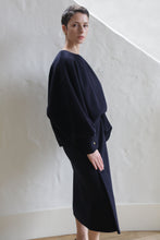 Load image into Gallery viewer, A person with short hair stands indoors against a textured white wall, wearing the Cocoon | Navy—a loose-fitting, dark navy long-sleeve top and matching skirt. They look at the camera with a neutral expression.