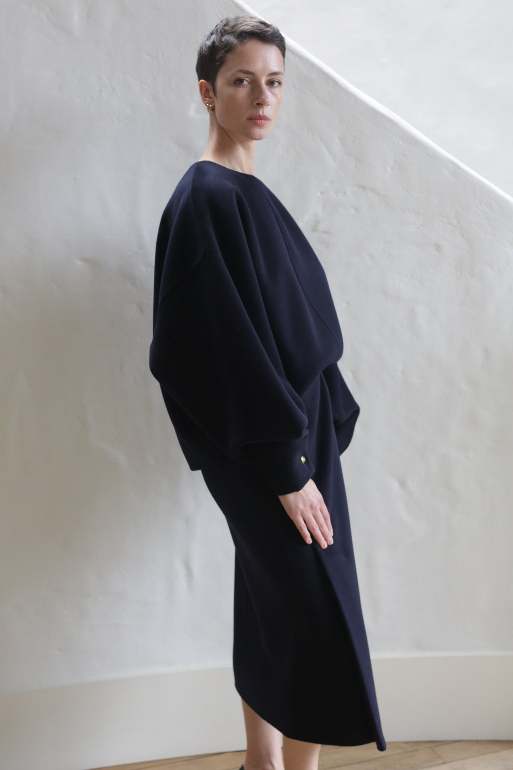 A woman with short hair stands indoors against a white textured wall, wearing the Wrap Skirt in navy. The skirt features a relaxed, loose fit and pairs with her long sleeves for a stylish, understated look.