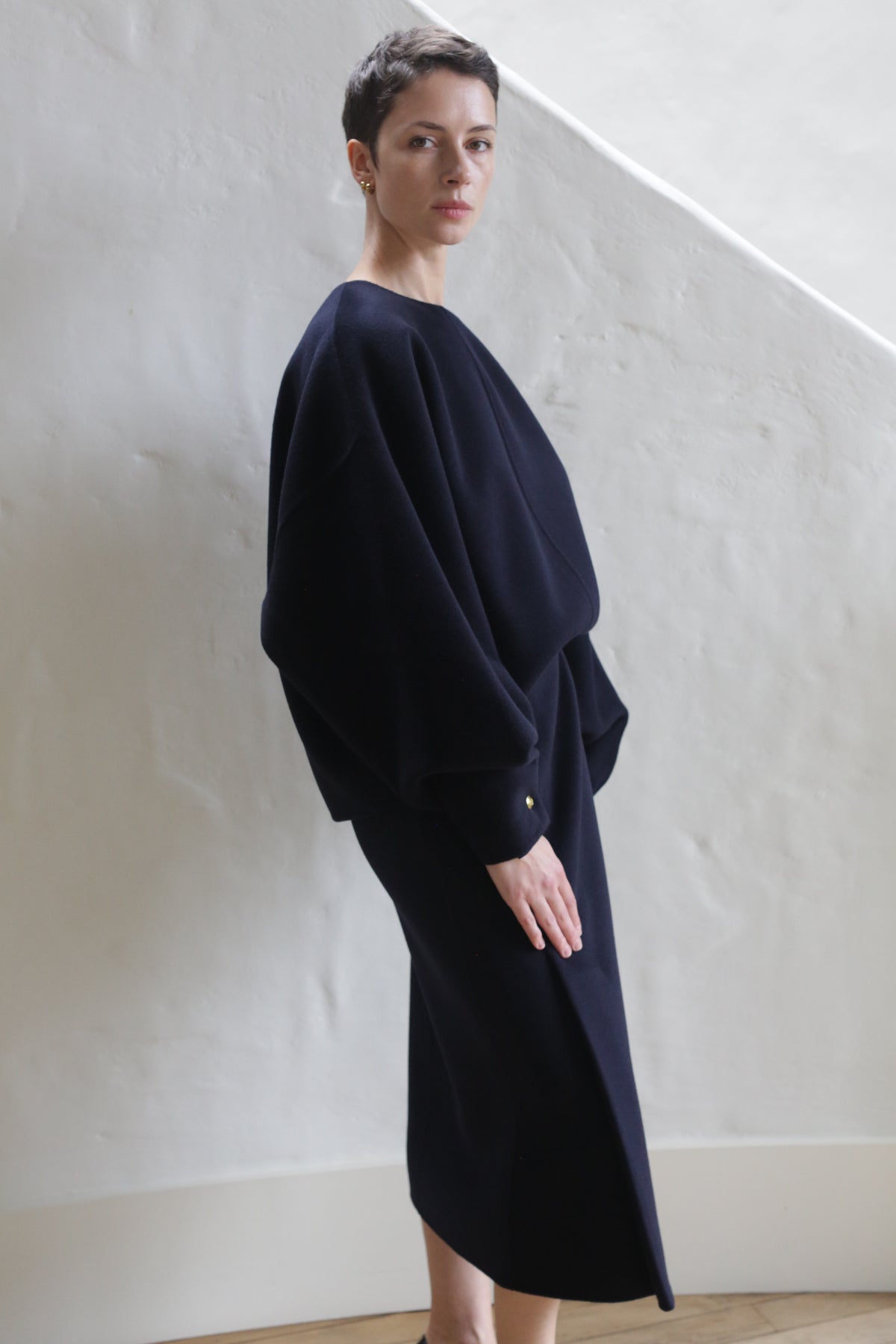 A woman with short hair stands indoors against a white textured wall, wearing the Wrap Skirt in navy. The skirt features a relaxed, loose fit and pairs with her long sleeves for a stylish, understated look.