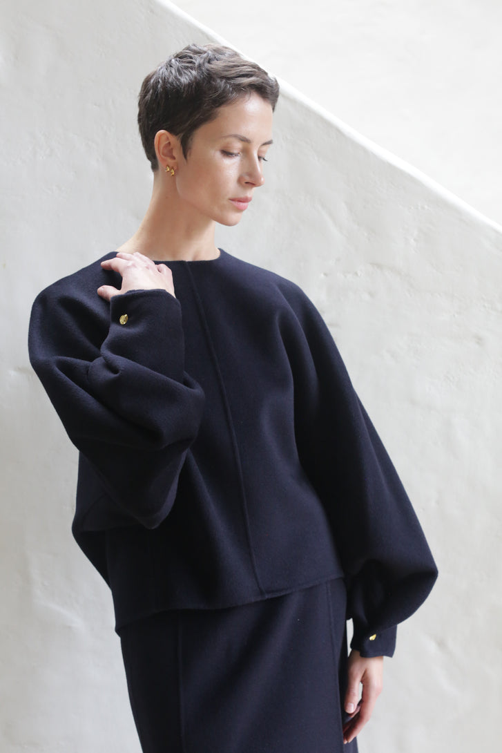 A woman with short hair models the Cocoon | Navy, a loose-fitting, long-sleeved top and matching skirt, standing against a white textured wall, looking down with one hand raised to her shoulder.