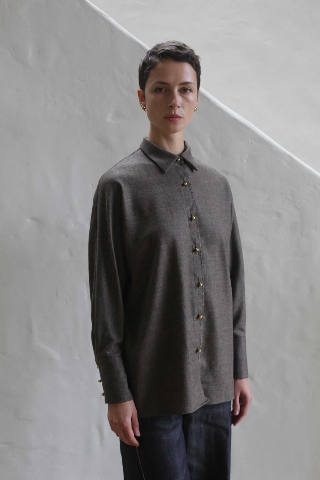 A person with short dark hair wears the Work Shirt | Taupe, a loose-fitting, long-sleeved button-up, standing indoors against a textured white wall by a slanted staircase.