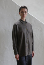 Load image into Gallery viewer, A person with short dark hair wears the Work Shirt | Taupe, a loose-fitting, long-sleeved button-up, standing indoors against a textured white wall by a slanted staircase.