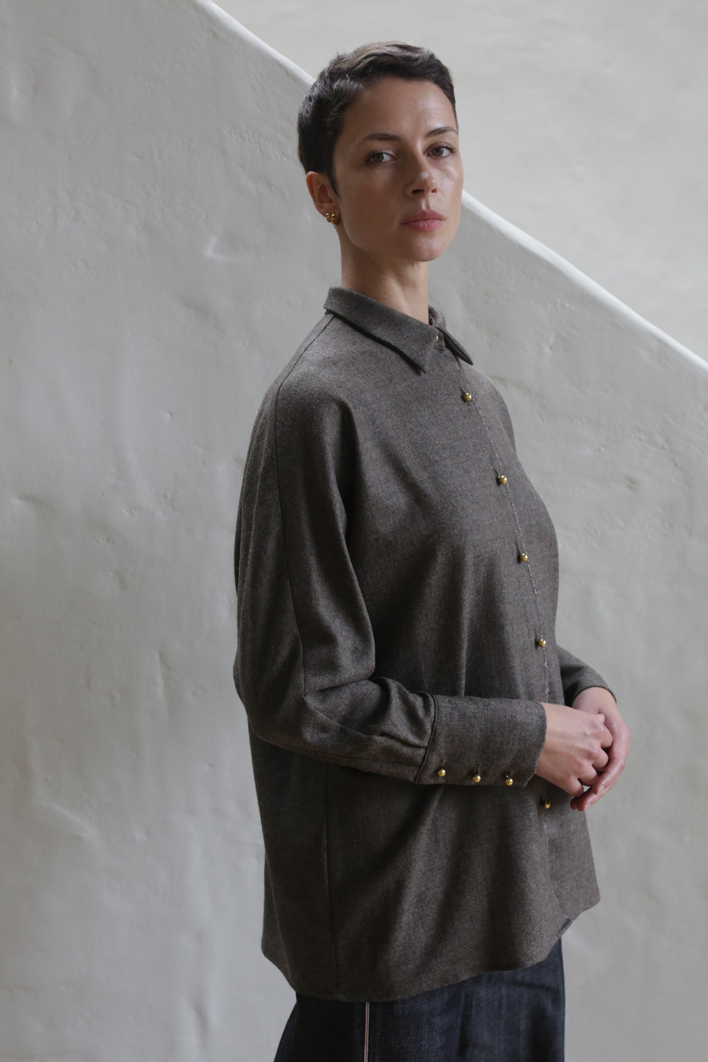 A woman with short brown hair stands in front of a light textured wall, wearing the Work Shirt | Taupe and dark pants, gazing slightly to the side with a neutral expression.