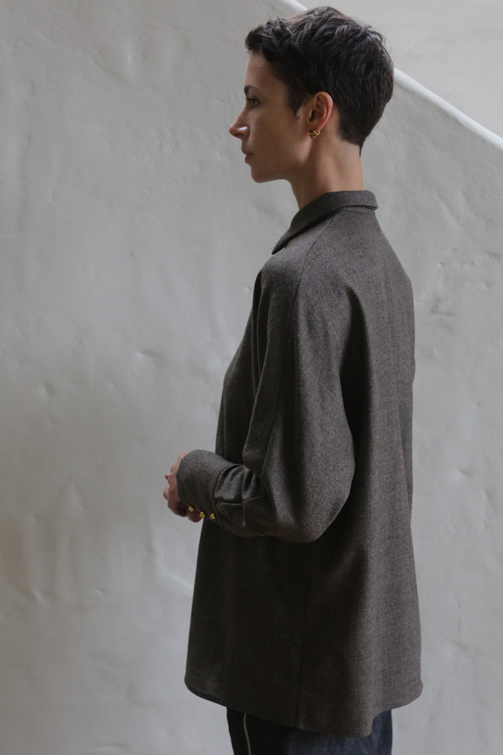A person with short dark hair, wearing the Work Shirt in Taupe, stands sideways against a light-colored textured wall and looks thoughtfully into the distance.