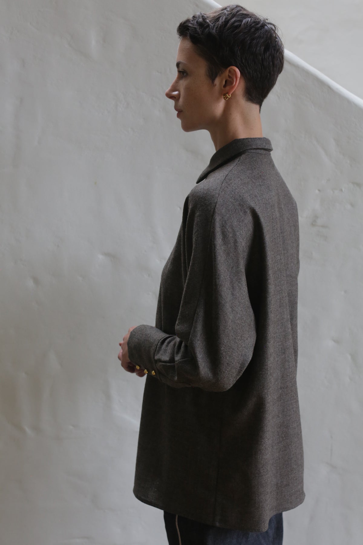 A person with short dark hair, wearing the Work Shirt in Taupe, stands sideways against a light-colored textured wall and looks thoughtfully into the distance.