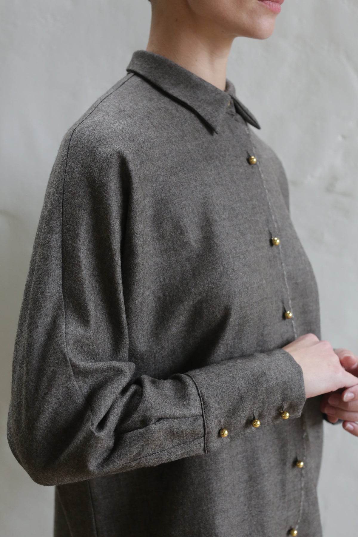 Someone stands against a light, textured background wearing the Work Shirt | Taupe. The gray button-up features gold buttons. Their face is partially out of frame as they gently clasp their hands.