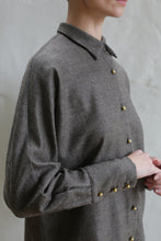 Load image into Gallery viewer, Someone stands against a light, textured background wearing the Work Shirt | Taupe. The gray button-up features gold buttons. Their face is partially out of frame as they gently clasp their hands.