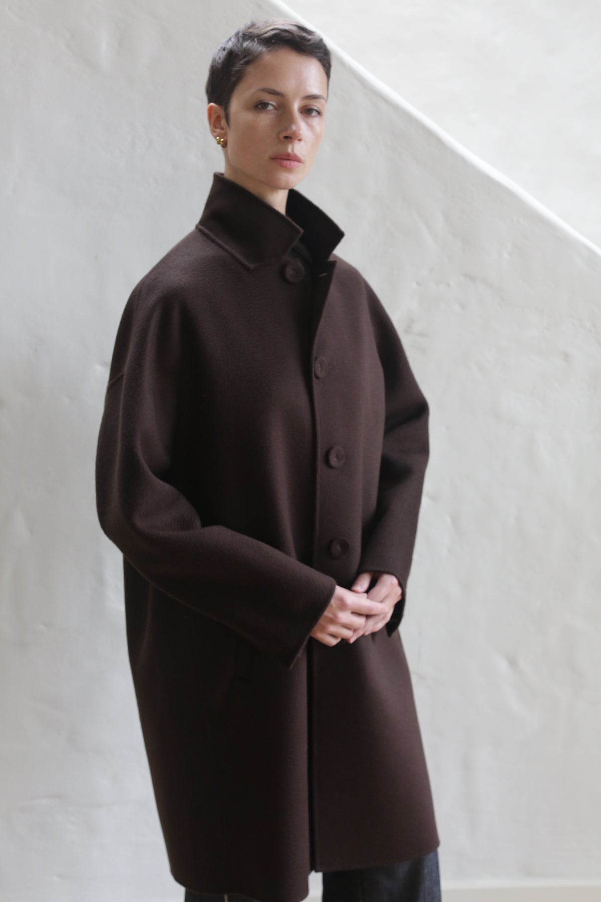 A person with short hair stands against a light wall, calmly facing the camera with hands clasped, dressed in the Men's Coat | Watermark Brown—a long, dark brown buttoned coat with a high collar.