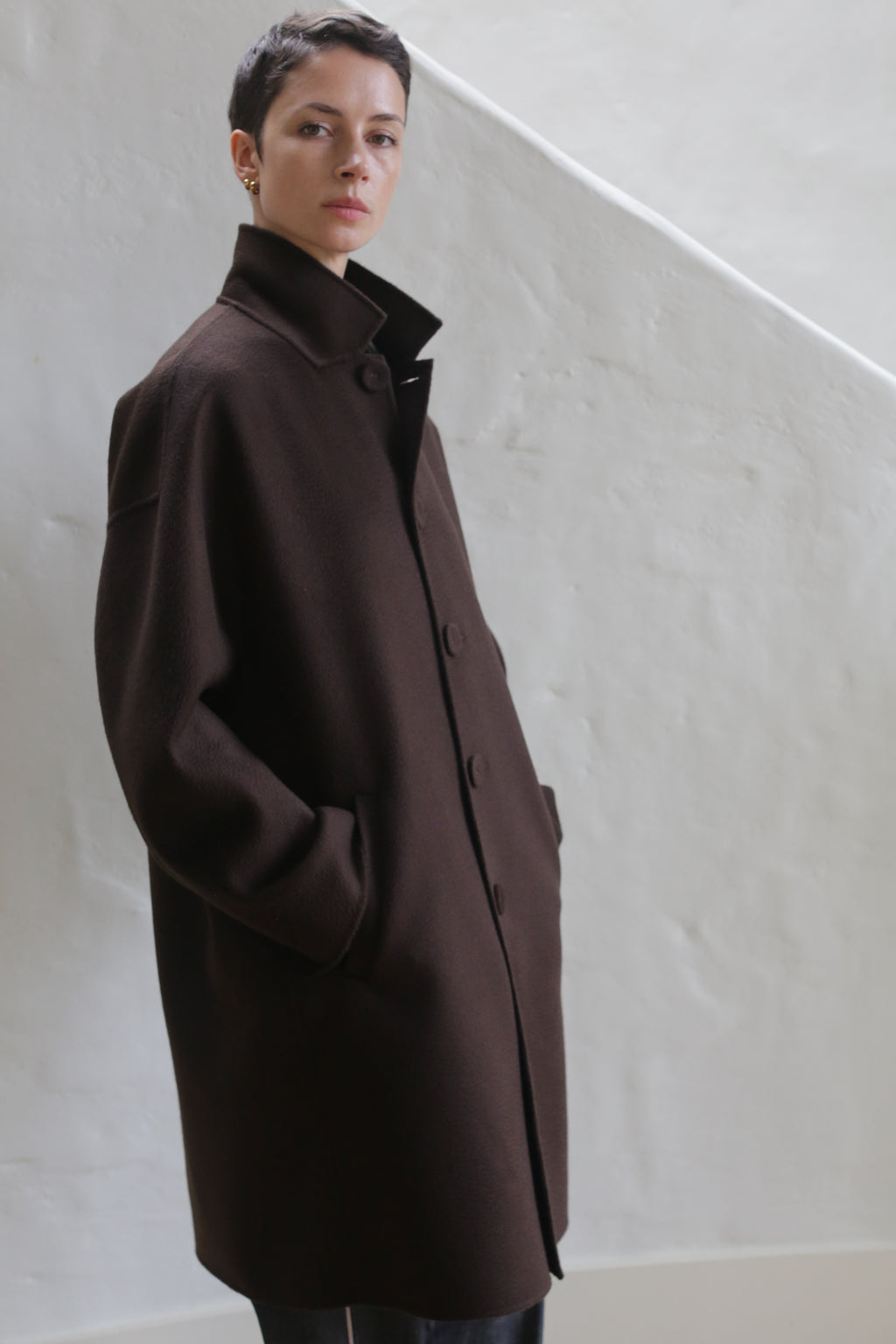 A man with short hair, hands in pockets, wears the Men's Coat | Watermark Brown while standing against a light textured wall with a diagonal architectural detail in the background.