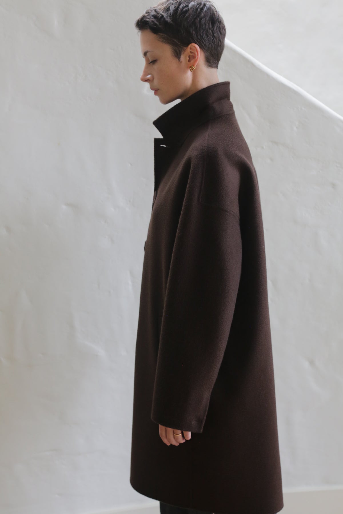 A man with short dark hair, dressed in the Men's Coat | Watermark Brown, stands in profile against a plain, light-colored wall.