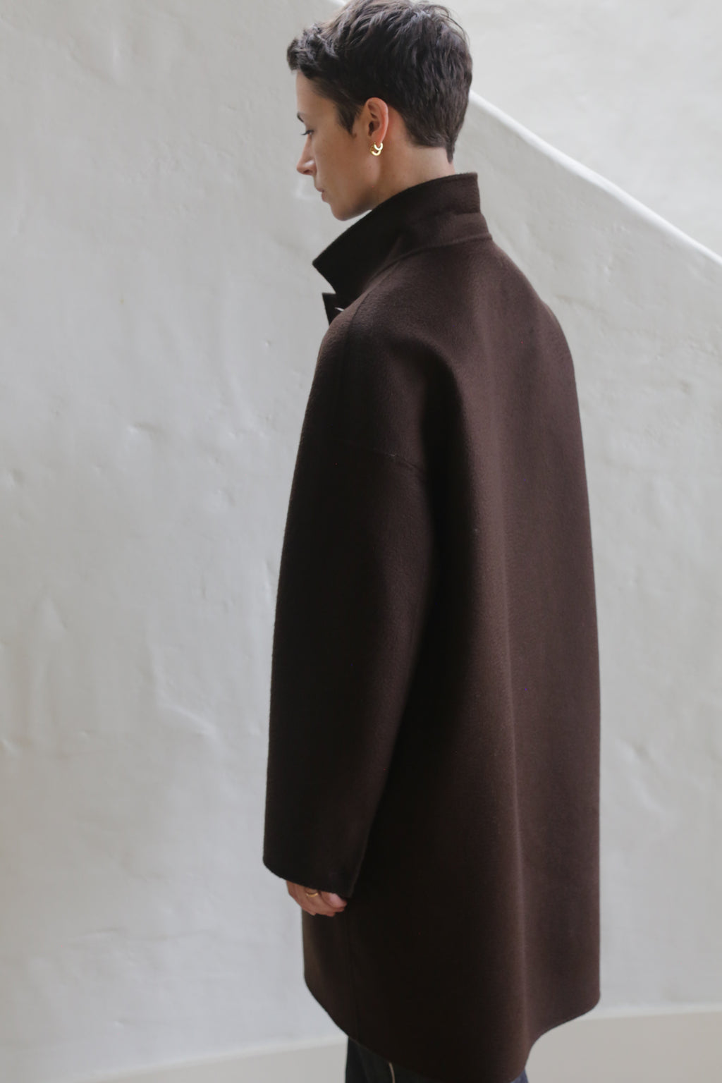 A person with short dark hair, wearing the Men's Coat | Watermark Brown, stands indoors facing away from the camera against a white textured wall.