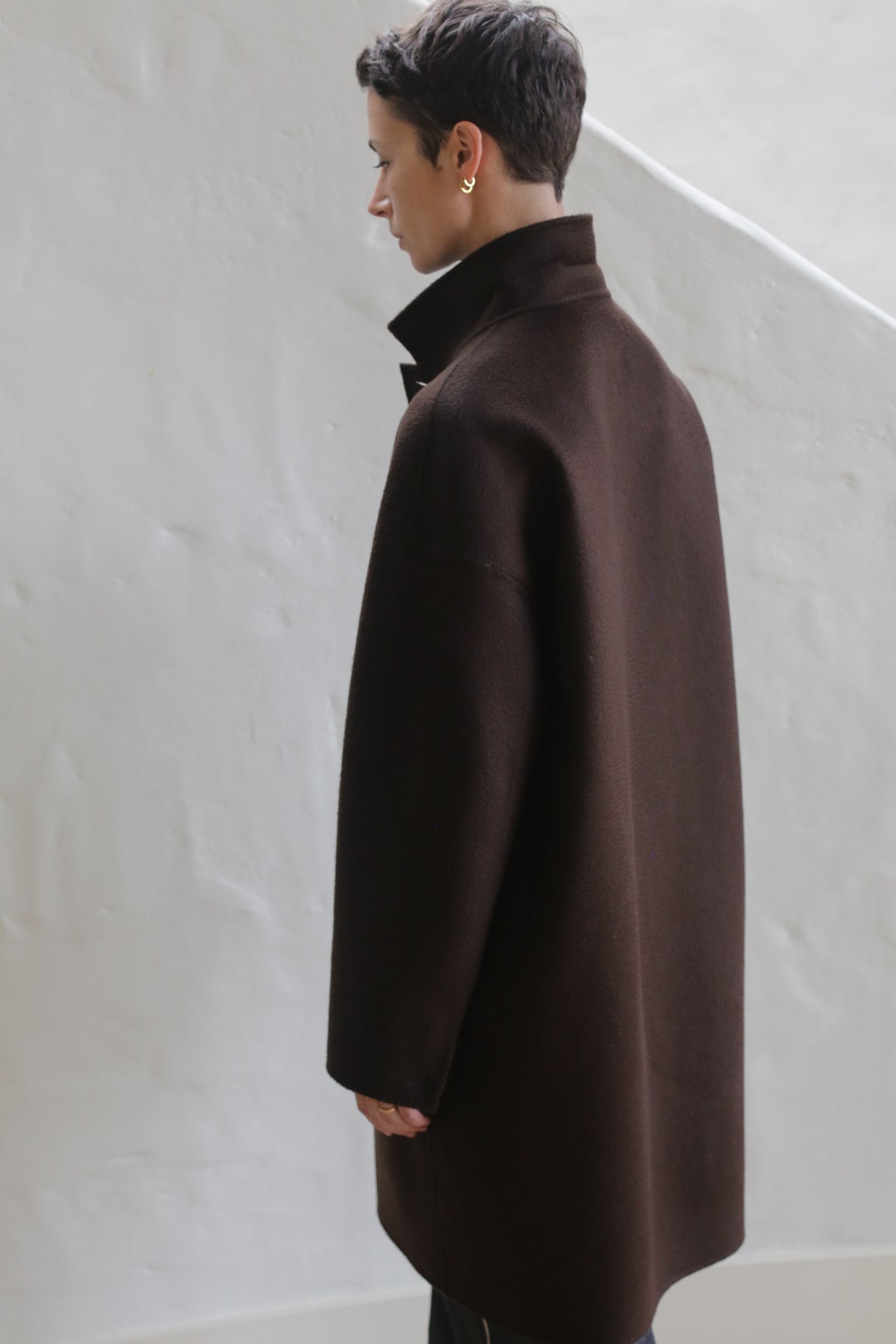 A person with short dark hair, wearing the Men's Coat | Watermark Brown, stands indoors facing away from the camera against a white textured wall.