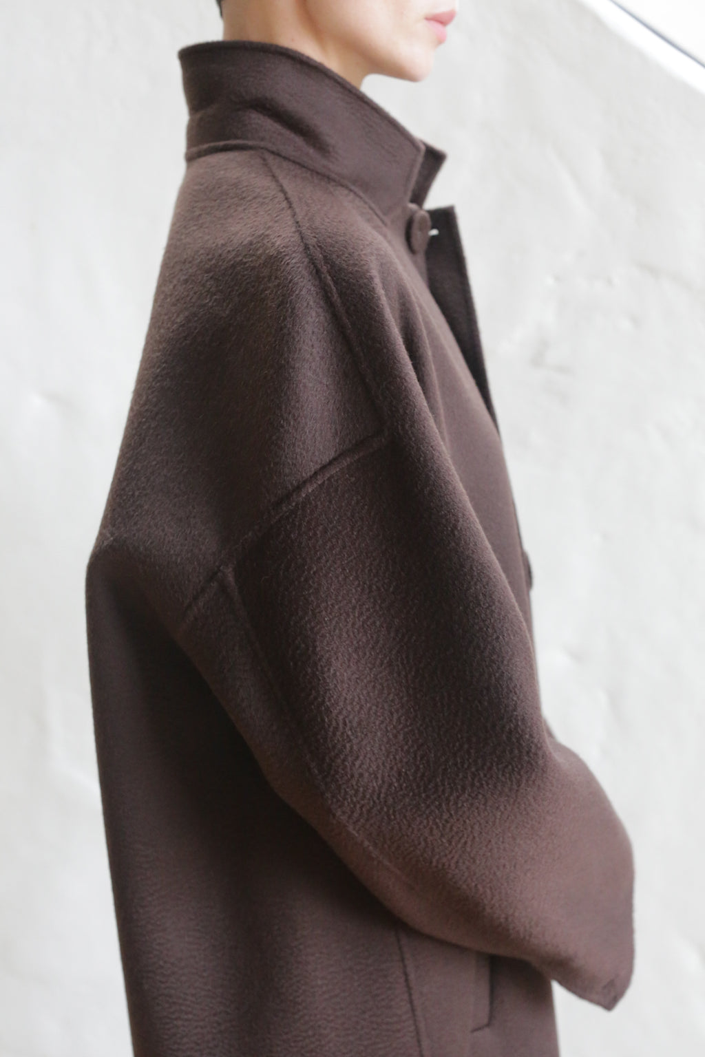 A person models the Men's Coat | Watermark Brown, its high collar and textured brown fabric highlighted against a soft, blurred background, with the focus on the coat’s details and structure rather than the face.