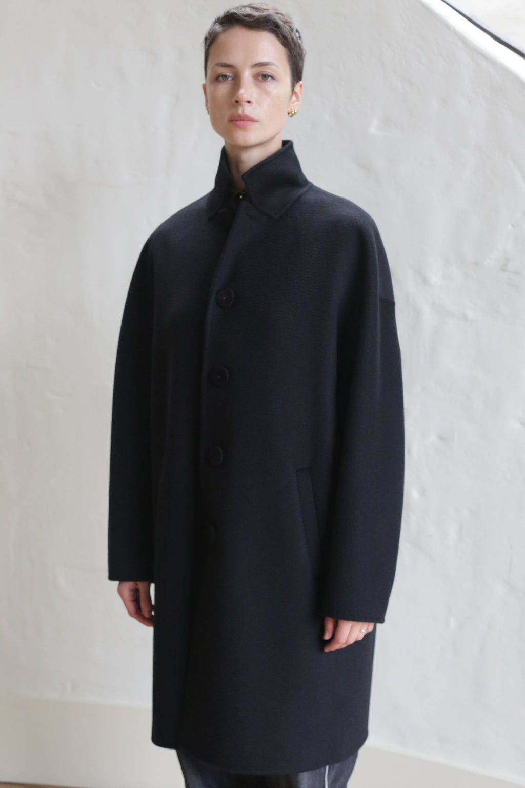 A person with short hair stands against a light, textured wall, wearing the Men's Coat | Watermark Black—a long, black, button-up coat featuring a high collar and side pockets.
