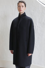 Load image into Gallery viewer, A person with short hair stands against a light, textured wall, wearing the Men&#39;s Coat | Watermark Black—a long, black, button-up coat featuring a high collar and side pockets.