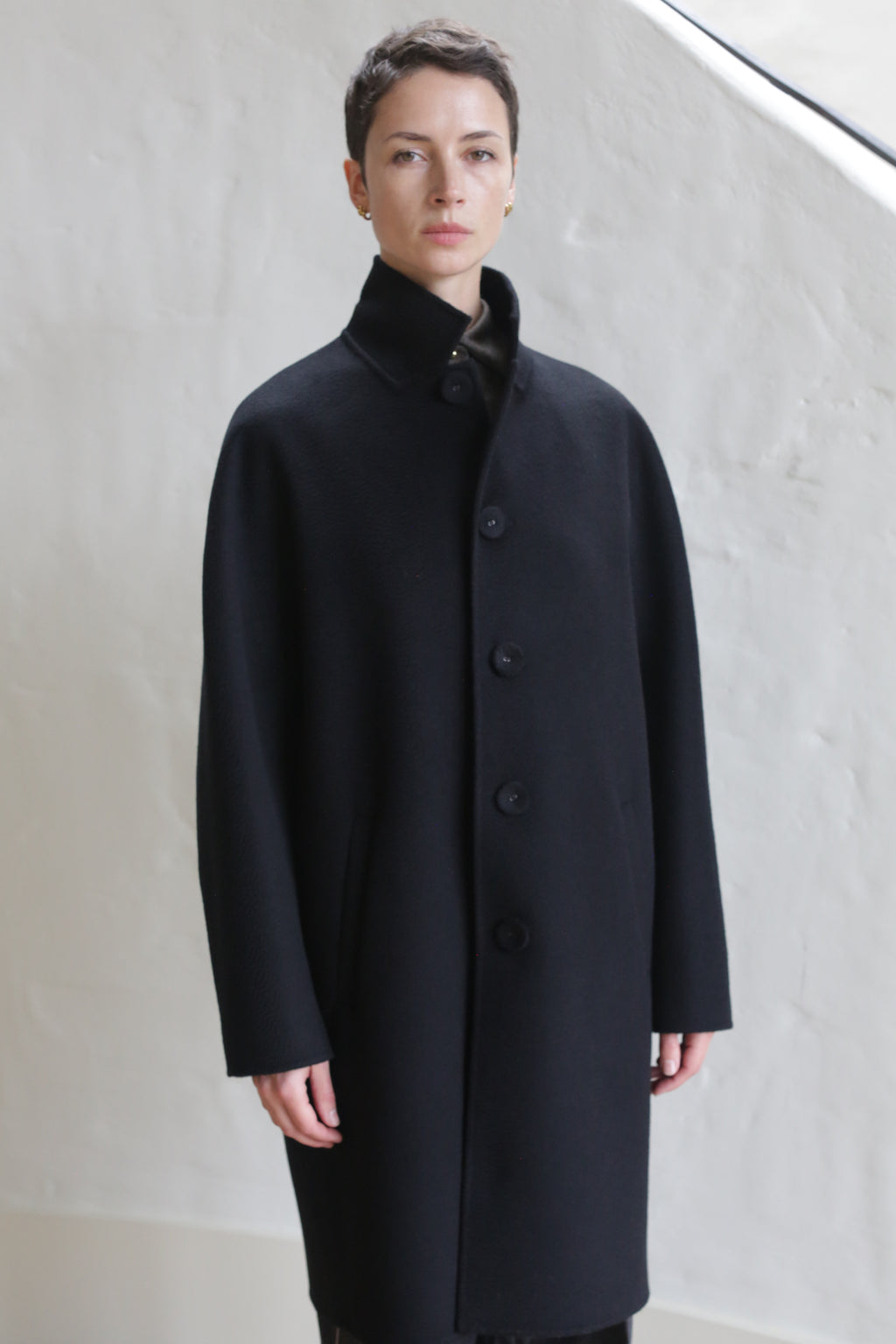 A man with short hair wears the Men’s Coat | Watermark Black, standing against a light-textured wall and looking directly at the camera with a neutral expression.