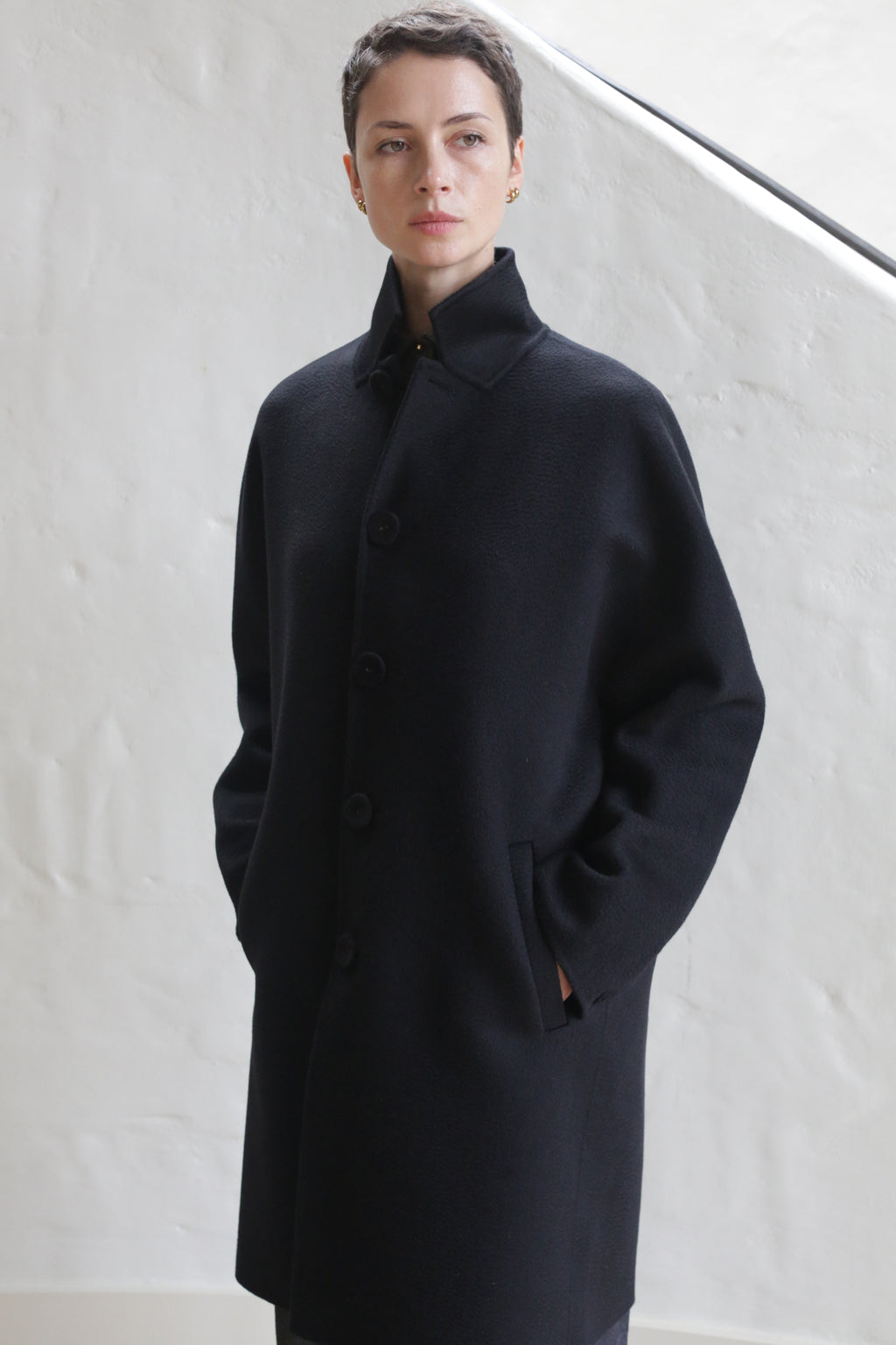 A person with short hair, hands in pockets, wears the Men's Coat | Watermark Black and stands against a white textured wall, gazing to the side with a neutral expression.