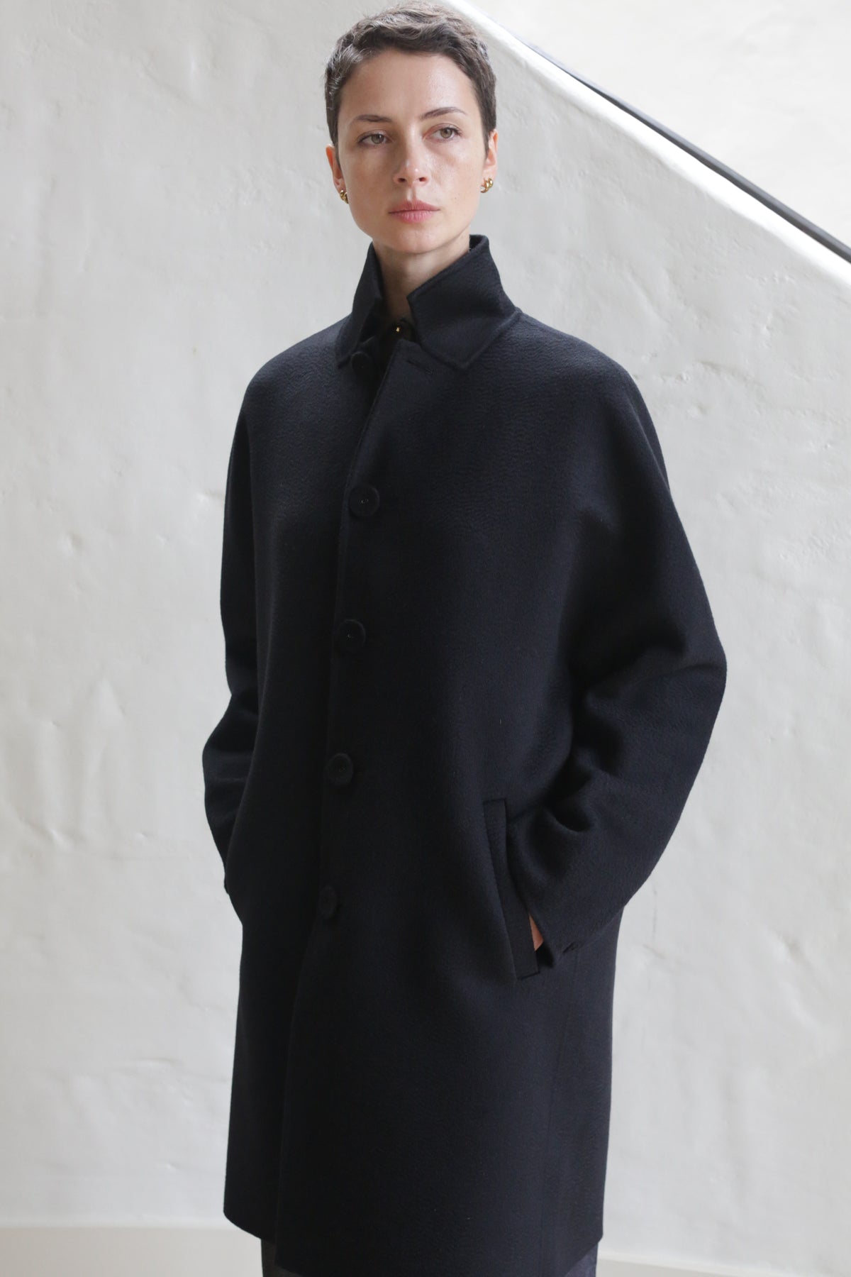 A person with short hair, hands in pockets, wears the Men's Coat | Watermark Black and stands against a white textured wall, gazing to the side with a neutral expression.