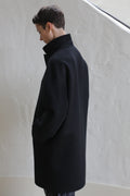 A person with short dark hair stands sideways, hands in pockets, wearing the Men's Coat | Watermark Black. They look down against a plain light-colored wall with soft natural lighting.