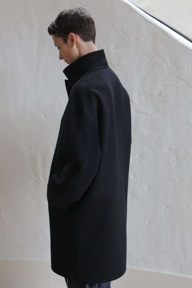 A person with short dark hair stands sideways, hands in pockets, wearing the Men's Coat | Watermark Black. They look down against a plain light-colored wall with soft natural lighting.