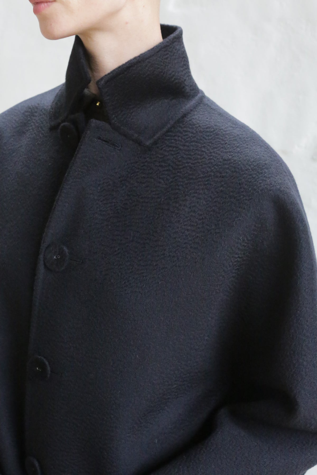 A man models the Men's Coat | Watermark Black, a dark, high-collared, textured coat buttoned to the top, shown from the shoulders up against a light background.