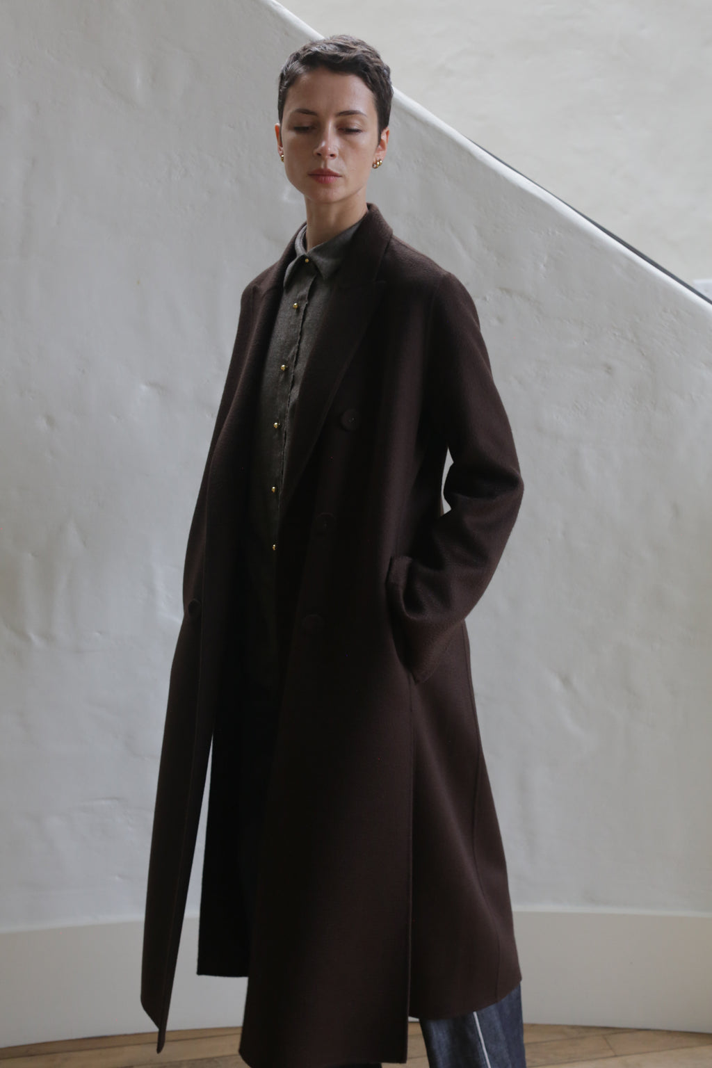 A person with short hair stands against a textured white wall wearing the Doublebreasted Coat in Watermark Brown over a button-up shirt and pants, hands in pockets, gazing slightly downward.
